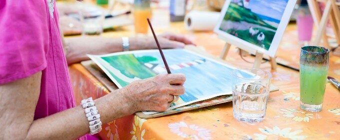 An older woman participates in an arts group activity, painting a picture of a lighthouse