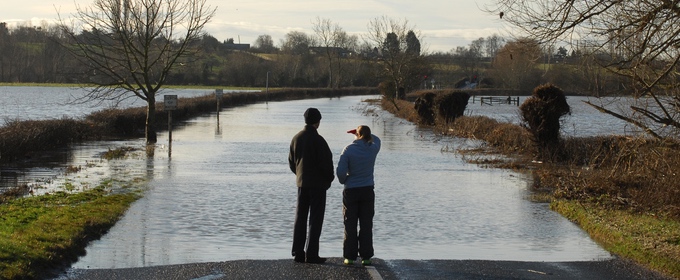 two adults standing at the edge of a flooded road in UK countryside.