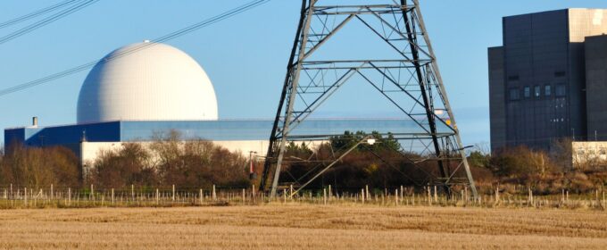 Image of a nuclear power station in the background with the bottom of an electricity pylon and a yellow grassy field in the foreground.