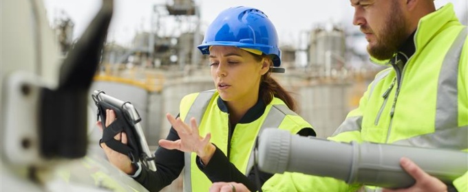 two workers with high-viz jackets and blue helmets are stood in a construction site looking at an I-pad