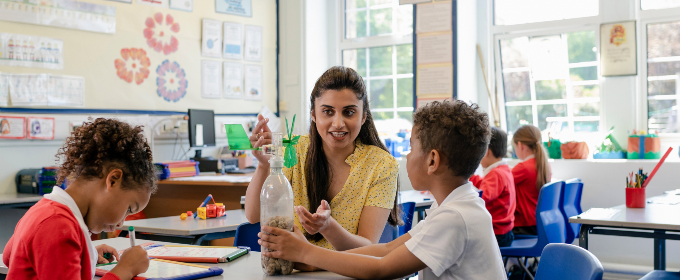 Teacher shows two students a science project using a plastic bottle in a bright, busy classroom.