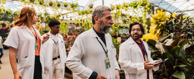 Doctors in white coats walking through a greenhouse. One talking and holding a clipboard.