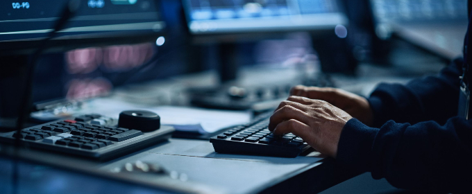 Photo of hands tapping at a computer keyboard, with multiple computer screens on the desk in front.