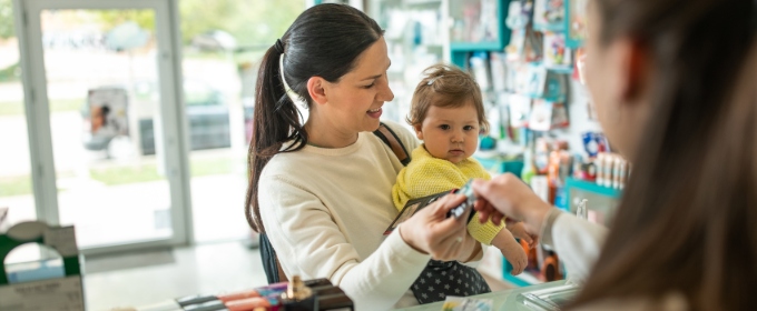 A mother holding an infant speaks over the counter to a pharmacy worker.