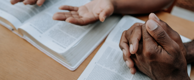 Close up image of hands, one pair clasped together, one pair open, over a religious text.